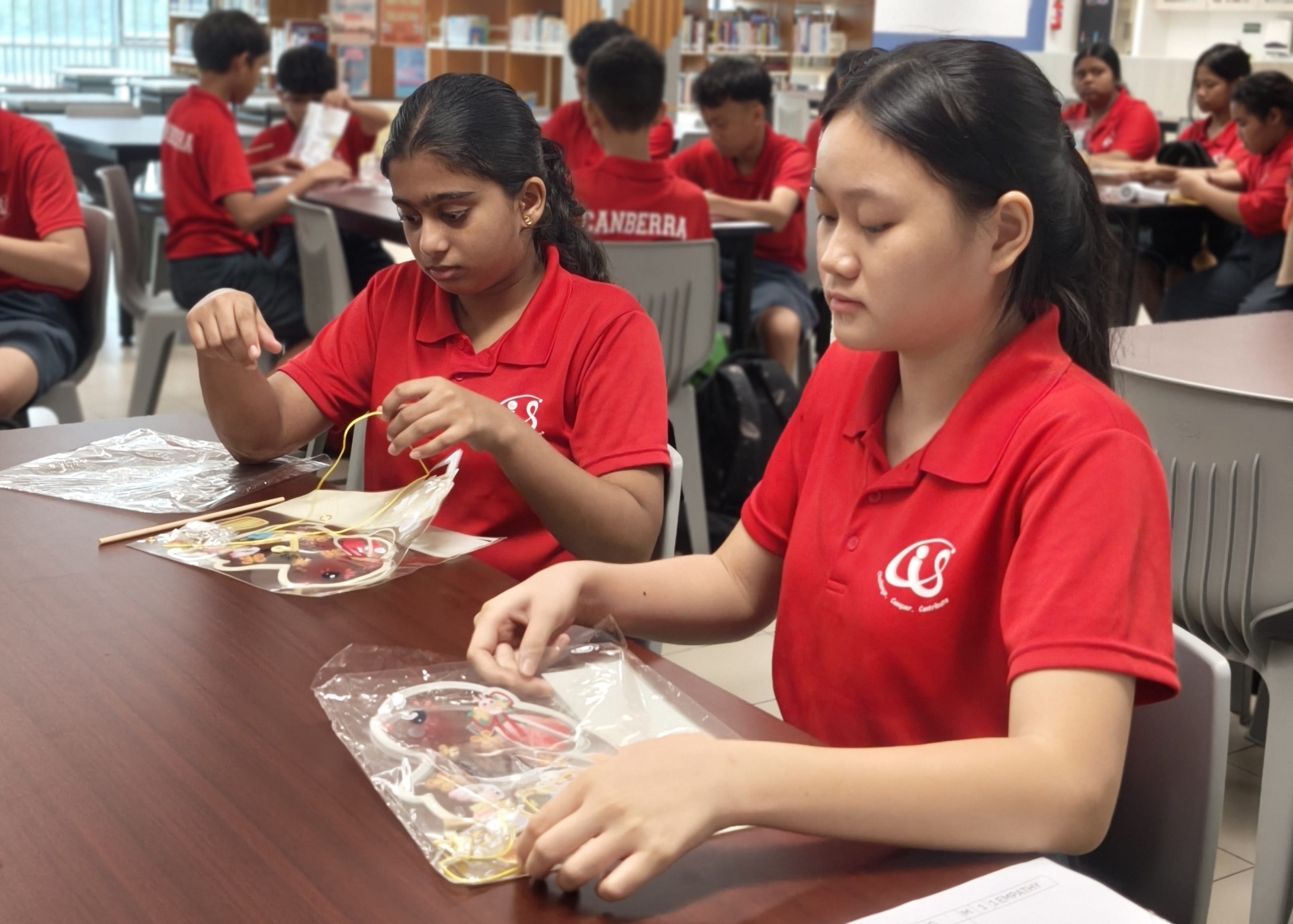 Two secondary school girls in red polo shirts sit at a table in a school library, carefully assembling their Mid-Autumn Festival lantern craft kits. Other students can be seen working on their lanterns in the background.